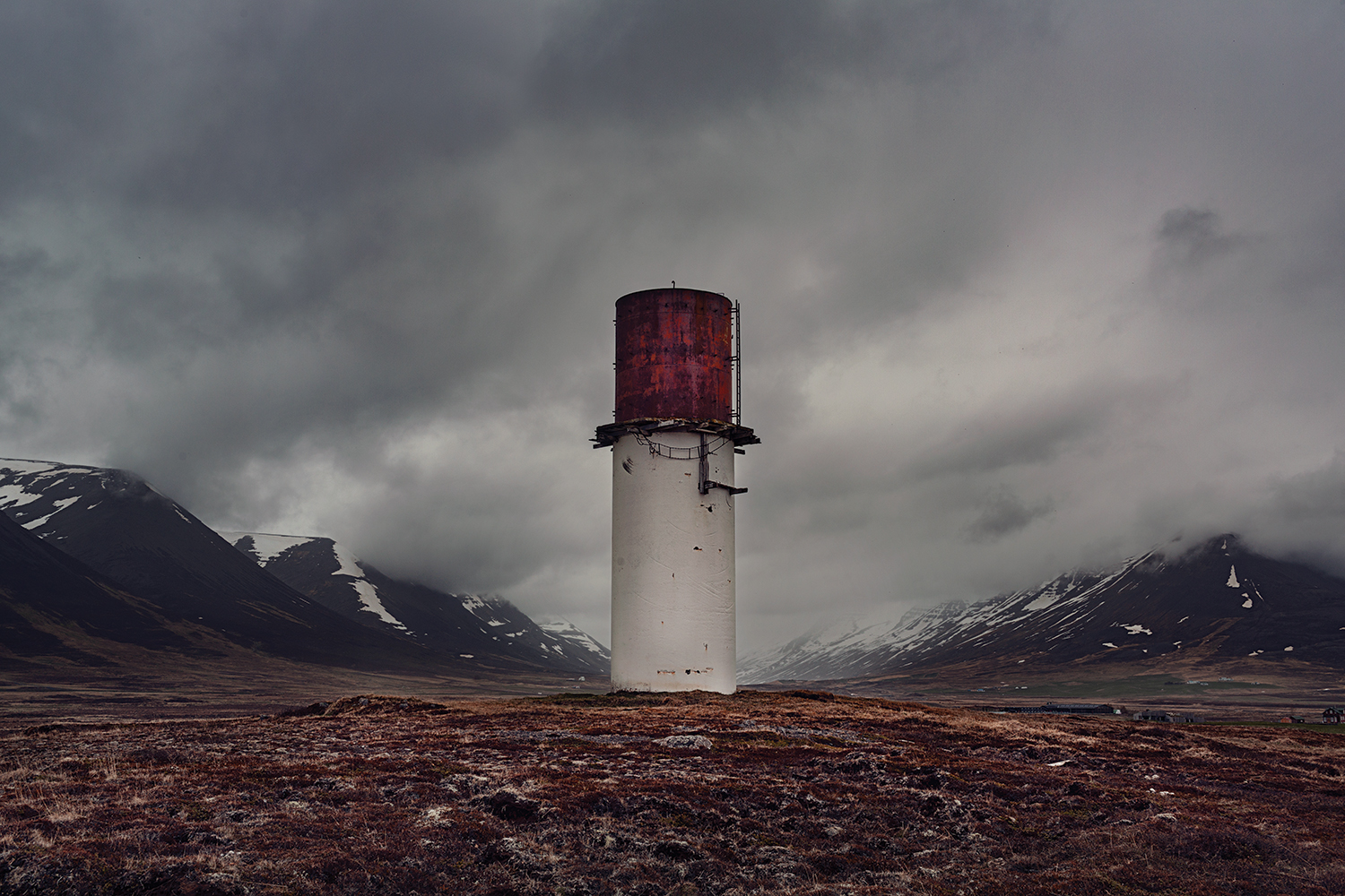 Alt description: An isolated rusted silo stands in an open field surrounded by mountains, with dark, heavy clouds overhead.