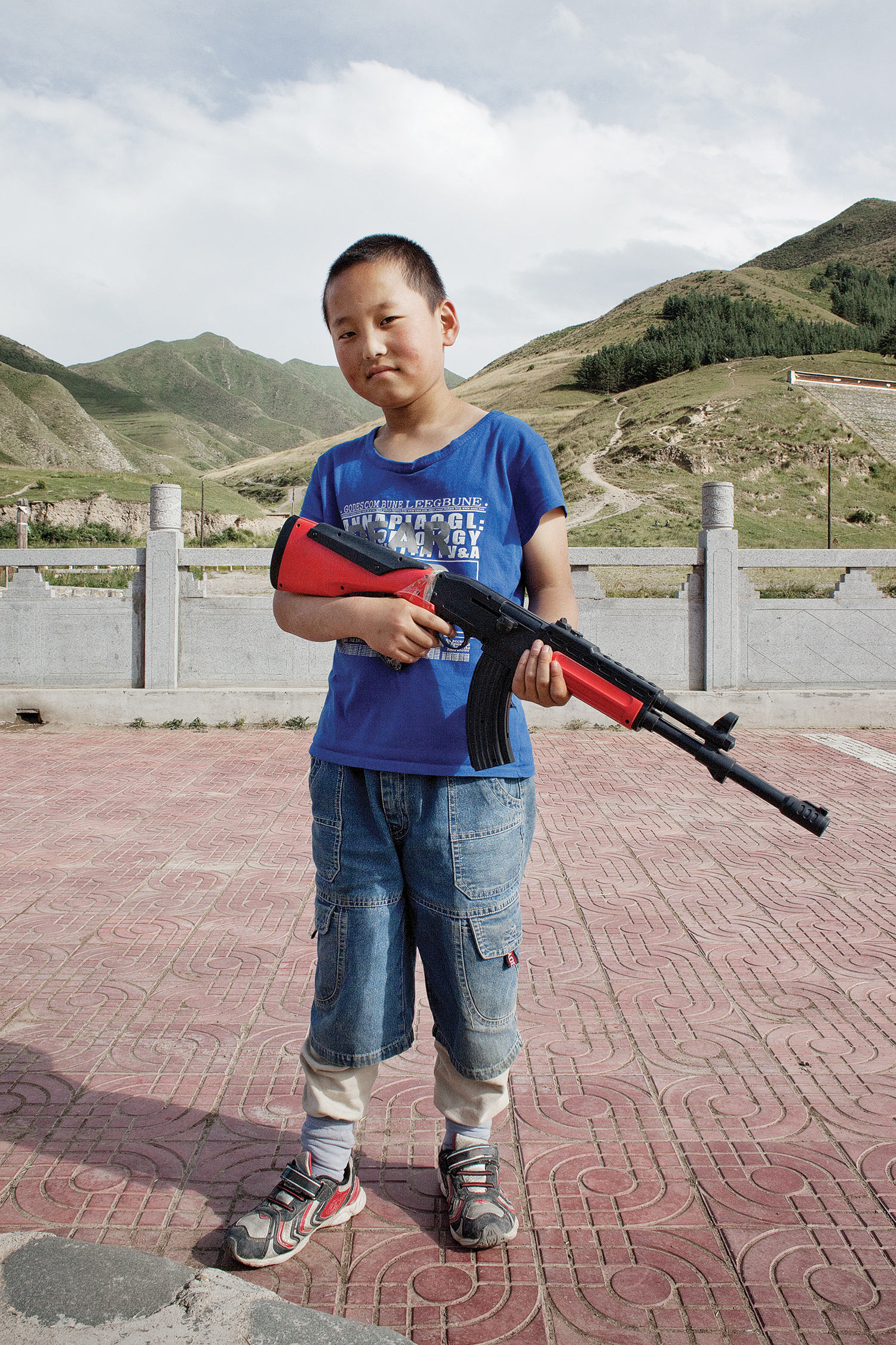 A young boy holding a toy gun poses confidently in an open area with rolling hills behind him in Xiahe County, Gansu Province, China. This image is by German photographer Florian Ritter.