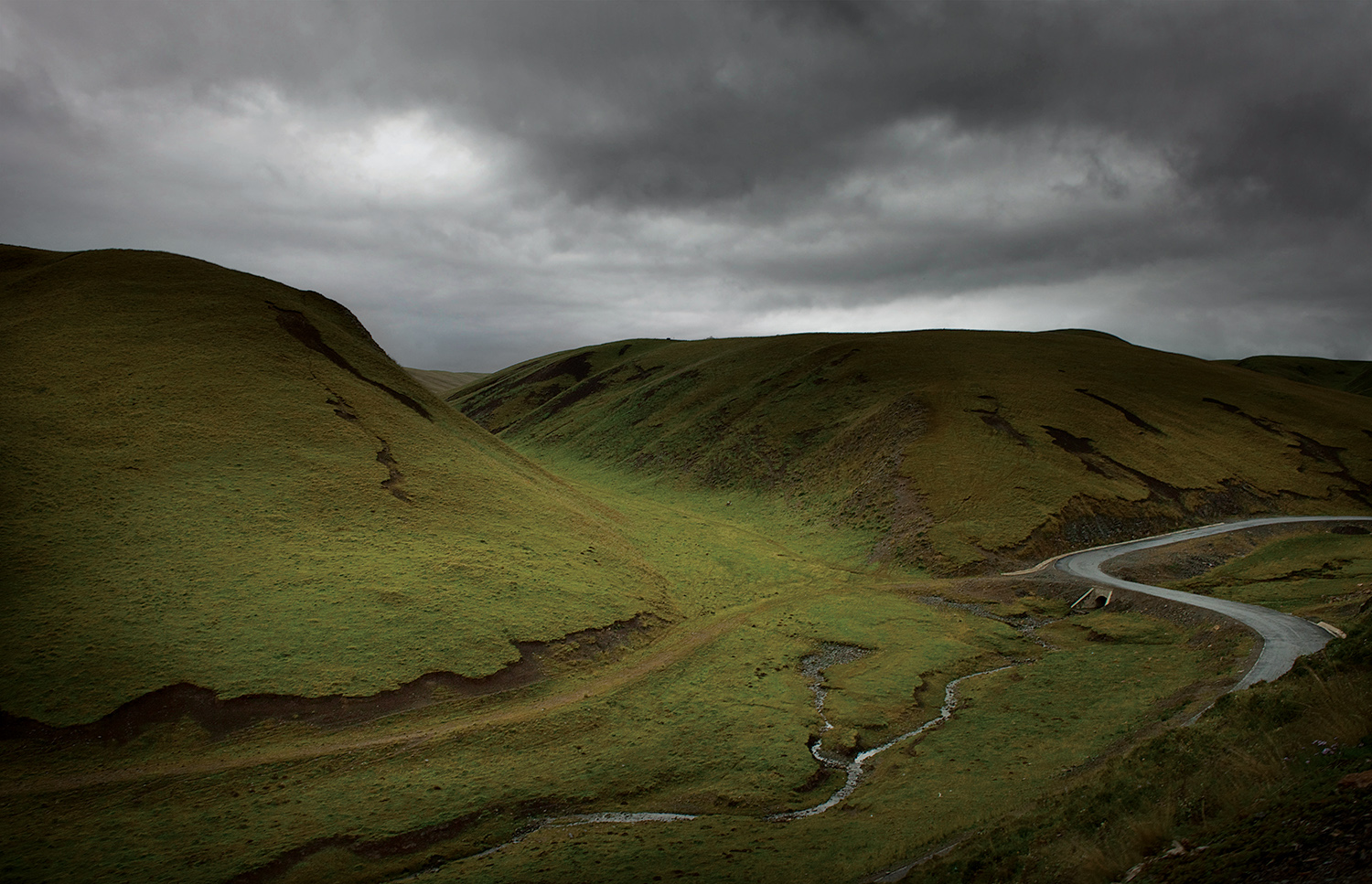 Scenic view of a winding road cutting through lush, rolling green hills under a stormy sky, photographed by Florian Ritter in Xiahe county, China.