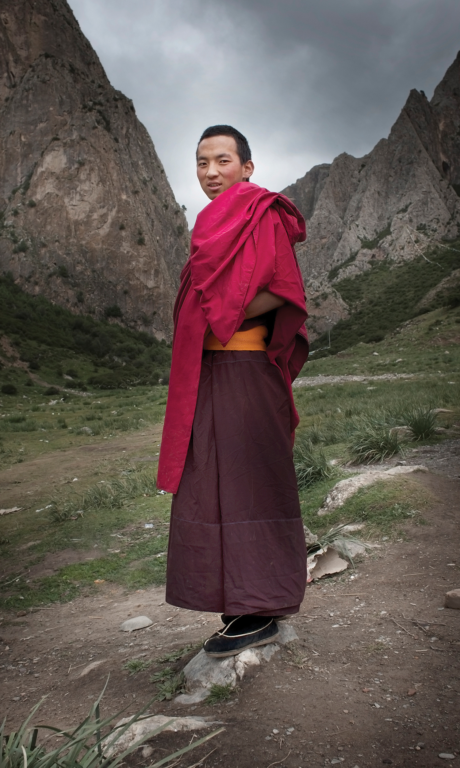 A Tibetan monk in red robes stands on a rocky path, with towering mountains behind him in Xiahe County, Gansu Province, China. This image is by German photographer Florian Ritter.